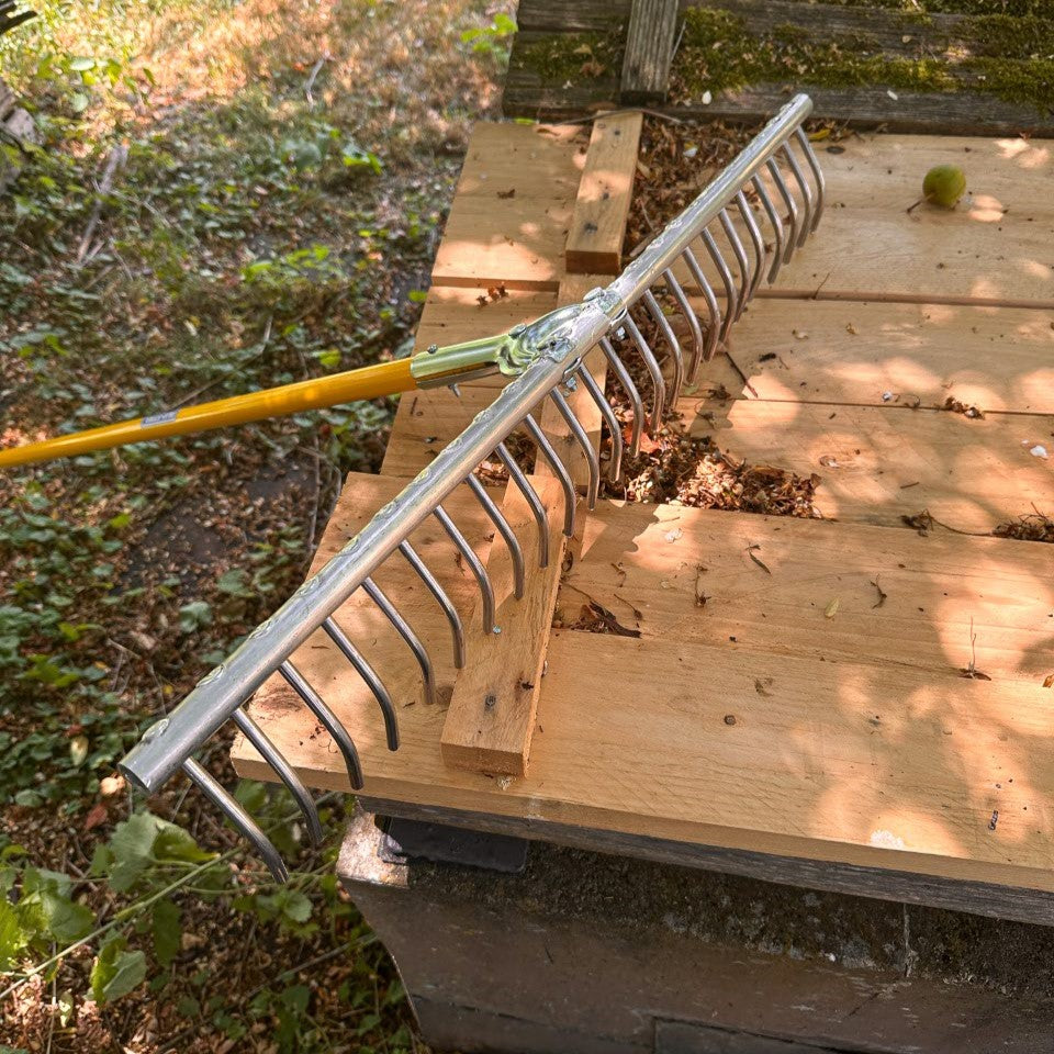 SHW Aluminium Heurechen mit silbernem Rechenkopf, gebogenen Zinken und montiertem gelbem Stiel auf einer Holzplattform im Garten. Detailansicht der Rechenkonstruktion.