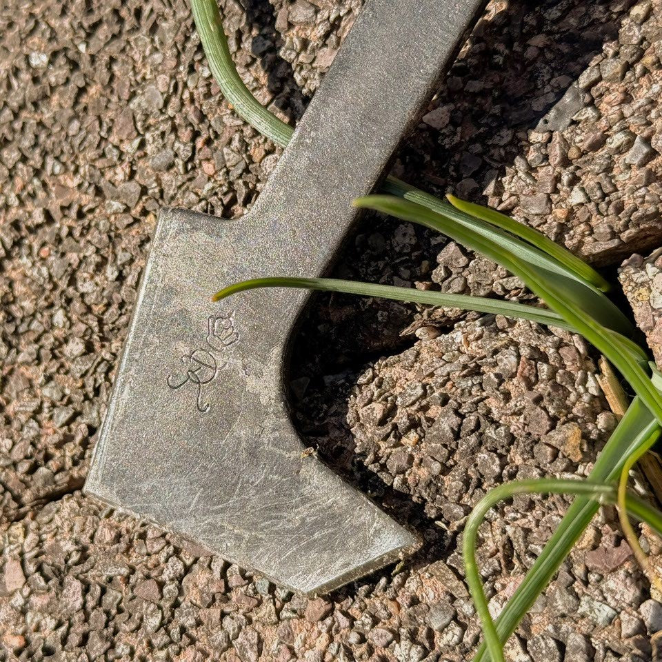 Nahaufnahme der abgewinkelten Stahlklinge des SHW Fugenkratzers mit eingeprägtem Herstellerzeichen auf einer Pflasterfläche. Die Klingenform eignet sich zum Auskratzen von Unkraut und Moos aus Fugen.