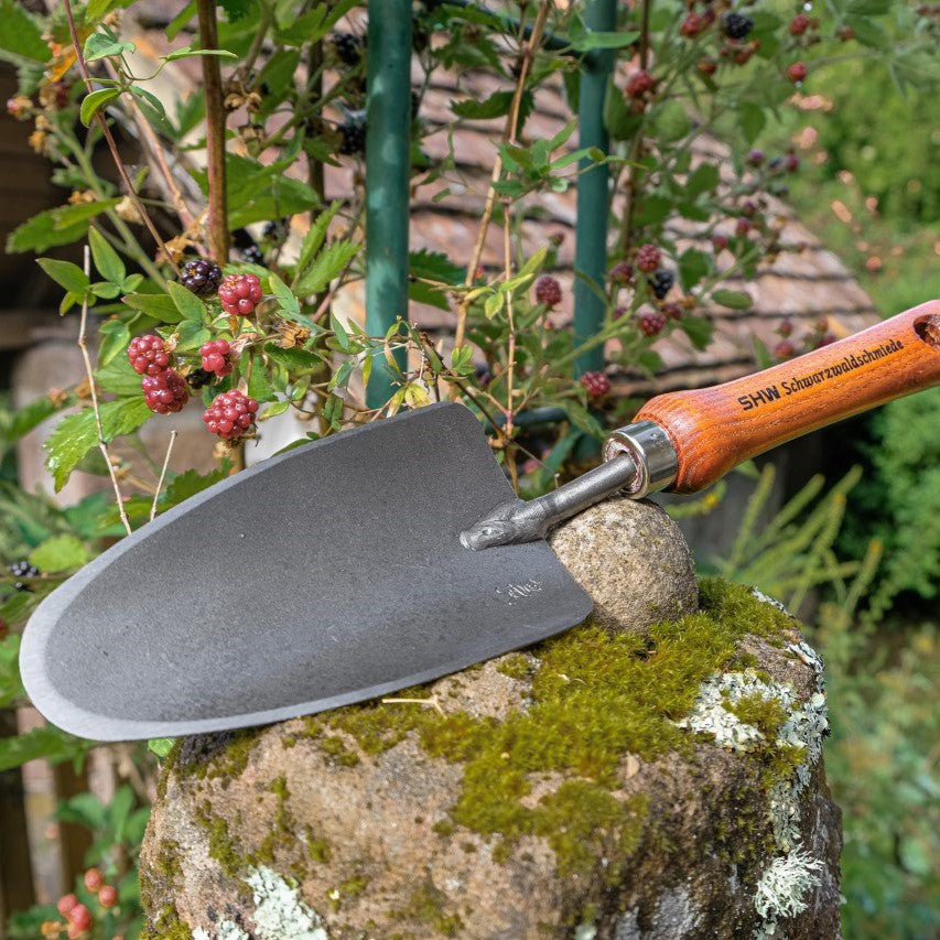 Nahaufnahme der breiten SHW Handschaufel mit geschmiedetem Stahlblatt und Holzgriff auf einem moosbewachsenen Natursteinpfosten neben einer Brombeerpflanze.