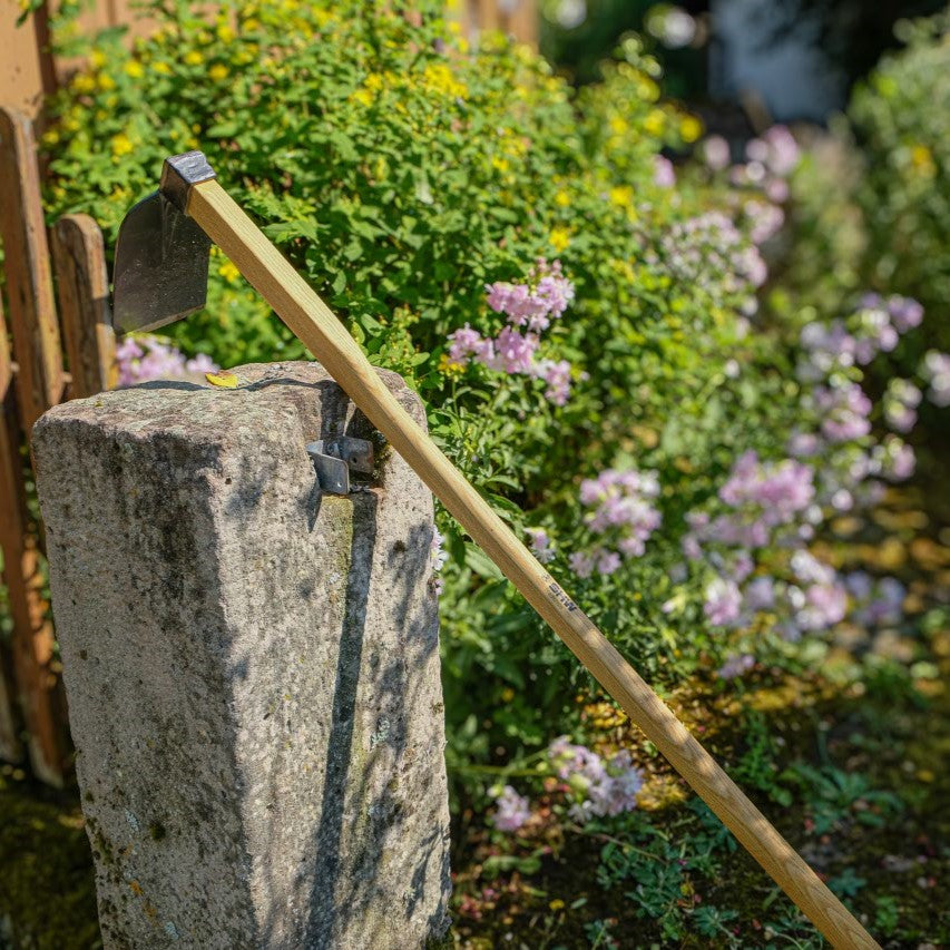 SHW Heilbronner Hacke mit breitem, geschmiedetem Hackenblatt und langem Holzstiel, angelehnt an einen Natursteinpfosten vor blühenden Gartenpflanzen. Für die Bodenbearbeitung im Garten.