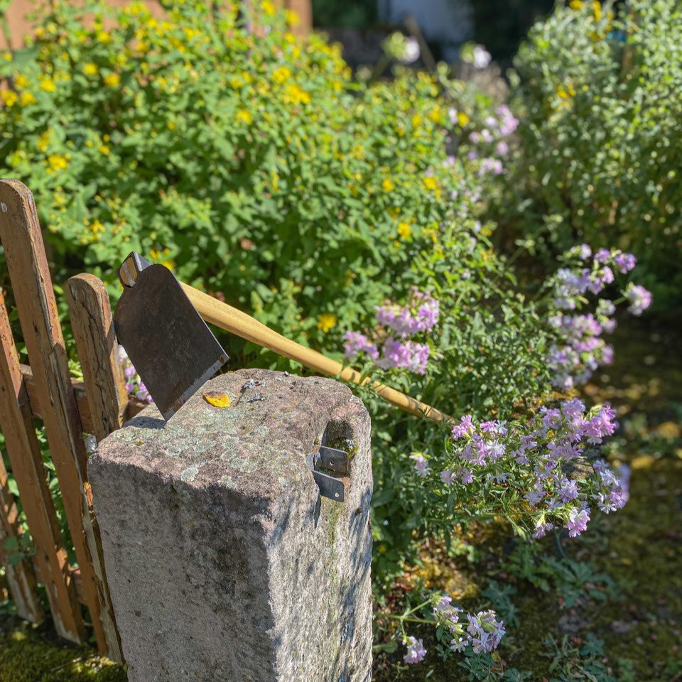 Nahaufnahme des breiten Stahlblatts der SHW Heilbronner Hacke mit Holzstiel auf einem Natursteinpfosten. Gartenhacke zum Hacken von Unkraut und Auflockern der Erde in Beeten.