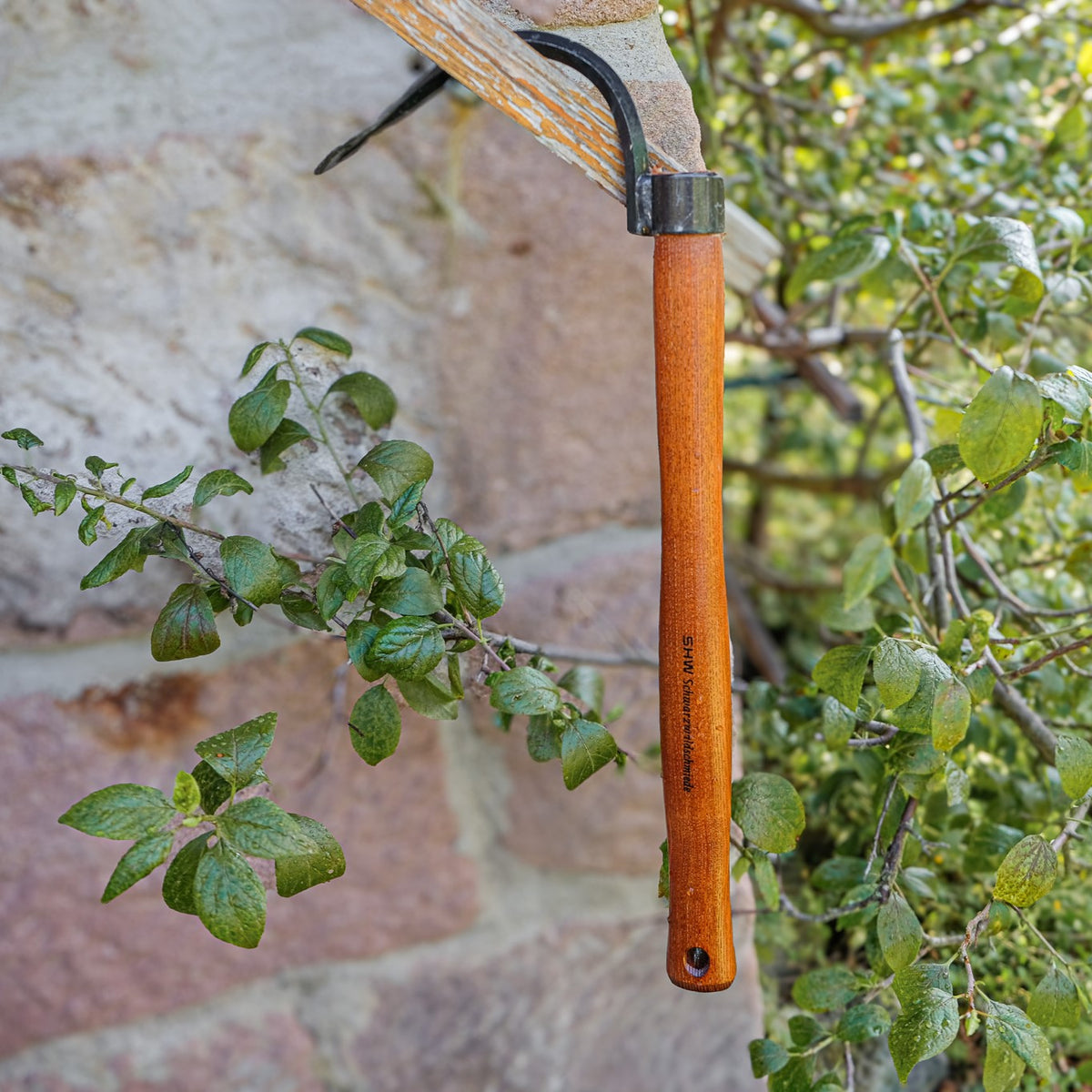 SHW Hochbeet Sauzahn mit braunem Holzgriff, gebogener Stahlzinke und spitzer Spitze, aufgehängt an einem Holzbalken zwischen grünen Gartenpflanzen vor einer Natursteinmauer.