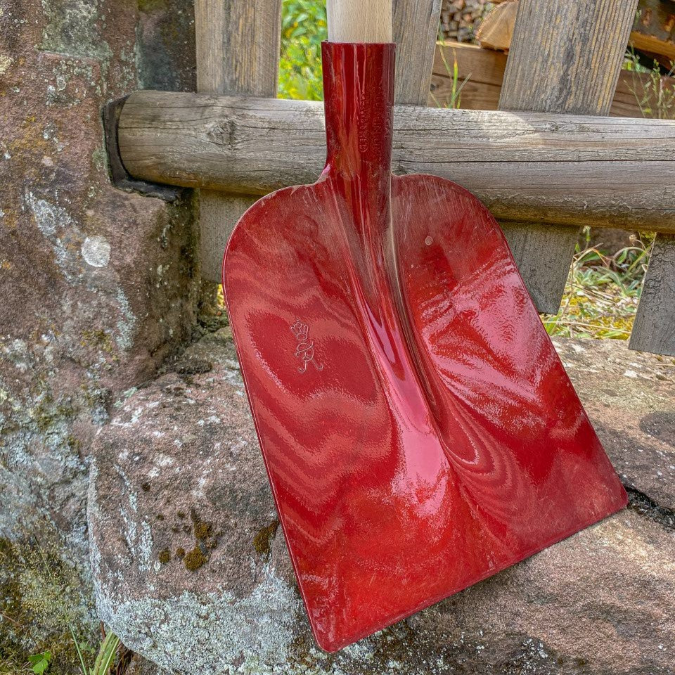 Nahaufnahme der roten SHW Holsteiner Aluminiumschaufel mit breitem Schaufelblatt, gerader Blattkante und Holzstiel, angelehnt an eine Natursteinmauer im Garten.