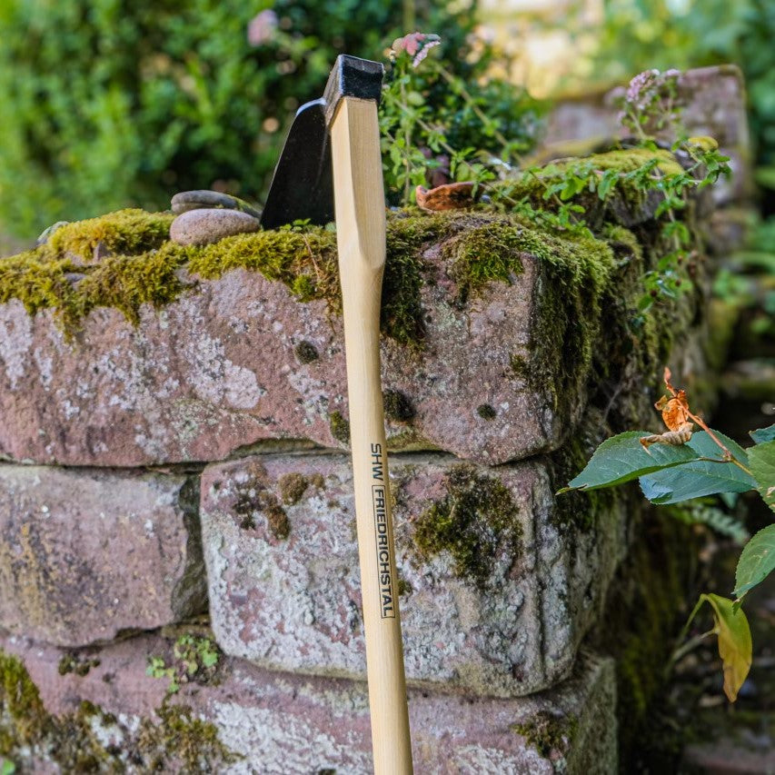SHW Reuthacke mit geschmiedetem Stahlkopf und ergonomisch geformtem Holzstiel vor einer moosbewachsenen Natursteinmauer. Robuste Gartenhacke zur Bodenbearbeitung und Entfernung von Unkraut.