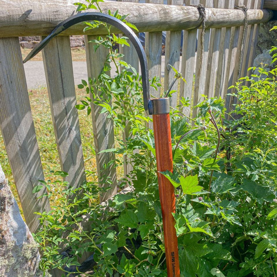 SHW Sauzahn mit langem Holzstiel und gebogener Stahlzinke, angelehnt an einen Holzzaun zwischen grünen Gartenpflanzen. Gartenwerkzeug zur tiefen Bodenlockerung ohne Umgraben.