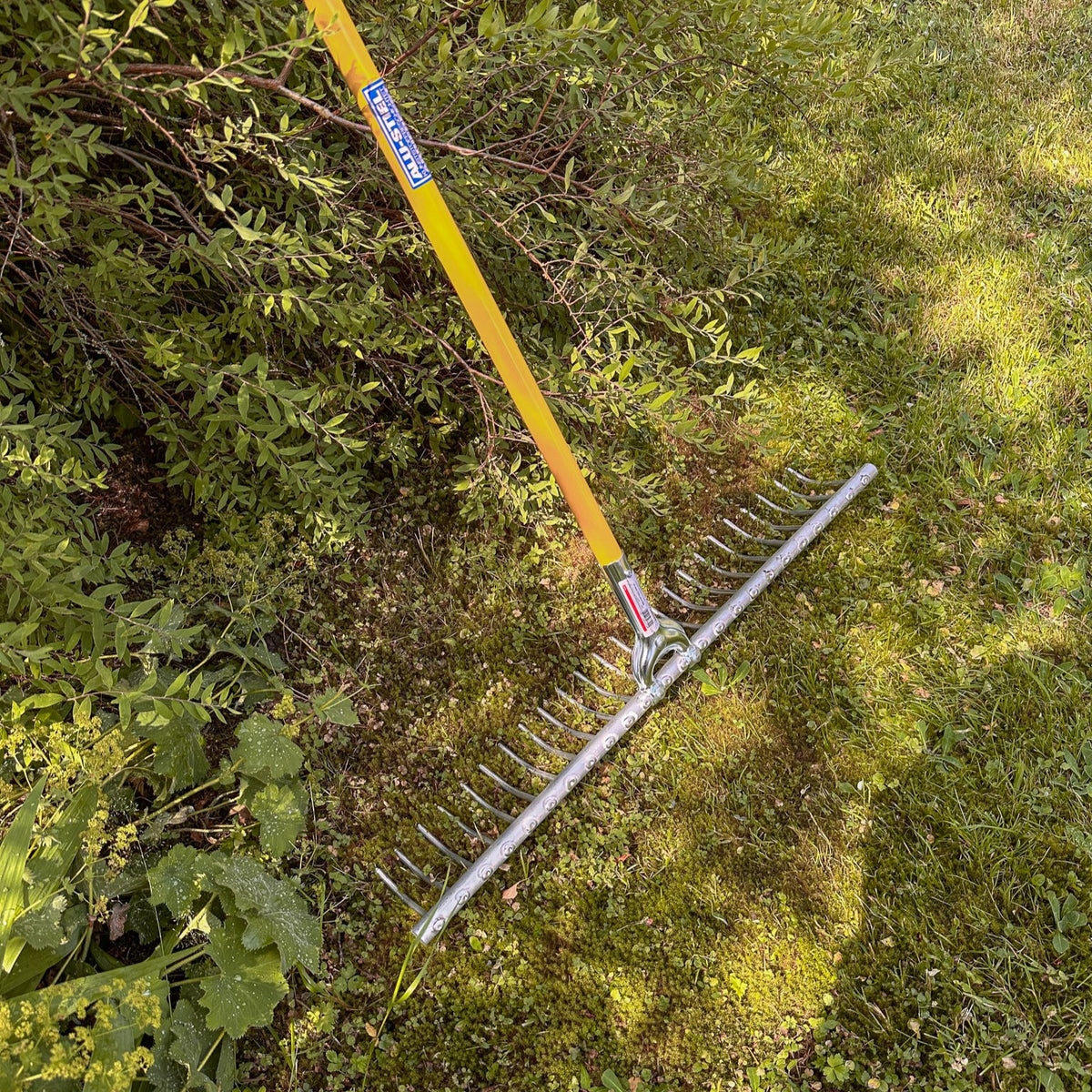 SHW Aluminium Heurechen mit gebogenen Metallzinken und montiertem gelbem Stiel auf einer moosbewachsenen Rasenfläche. Leichter Gartenrechen zum Zusammenrechen von Gras und Heu.