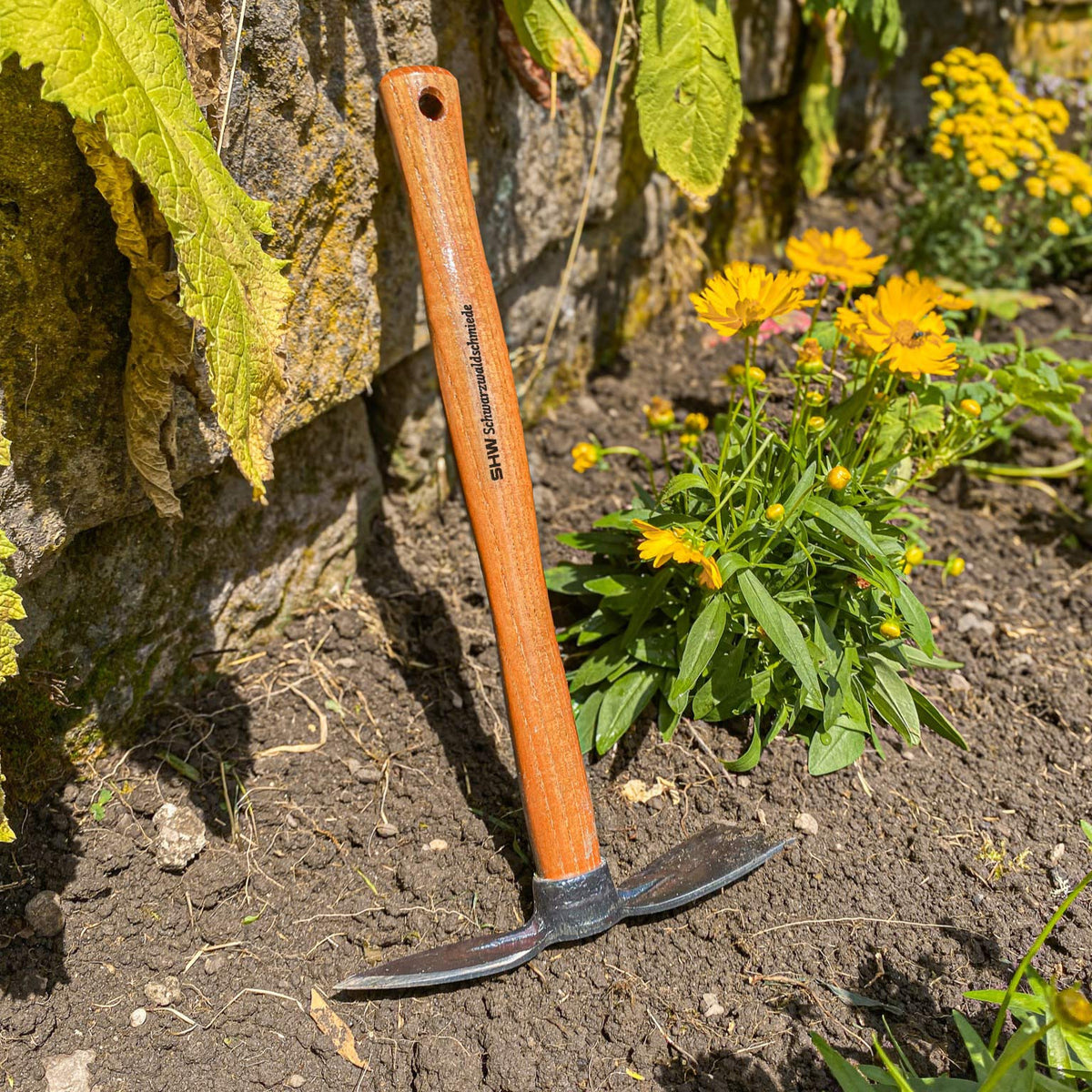SHW Haeckchenmit Holzstiel und geschmiedetem Stahlkopf, aufgestellt in einem Blumenbeet mit gelben Blüten. Robustes Gartenwerkzeug zum Lockern und Bearbeiten des Bodens.