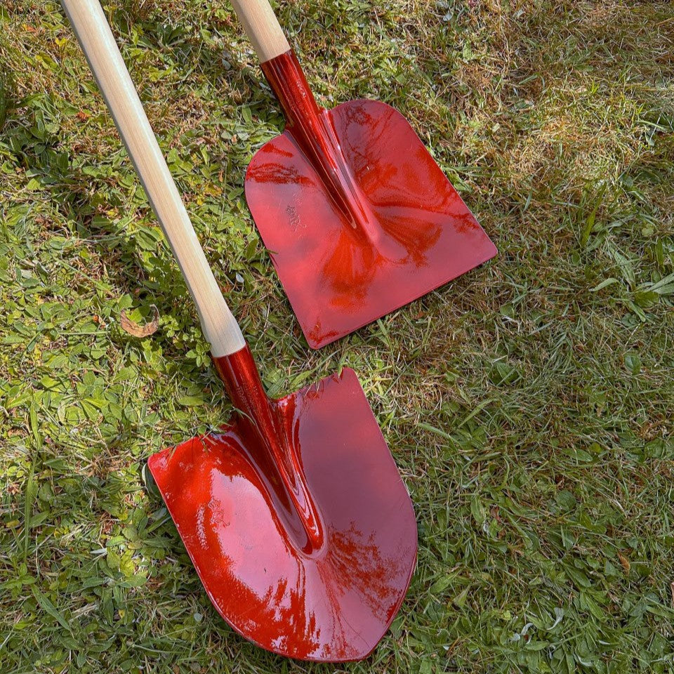 Zwei rote SHW Aluminiumschaufeln mit Holzstielen auf einer Rasenfläche. Vergleich zwischen der Holsteiner Schaufel mit gerader Blattkante und der Frankfurter Schaufel mit abgerundetem Schaufelblatt.