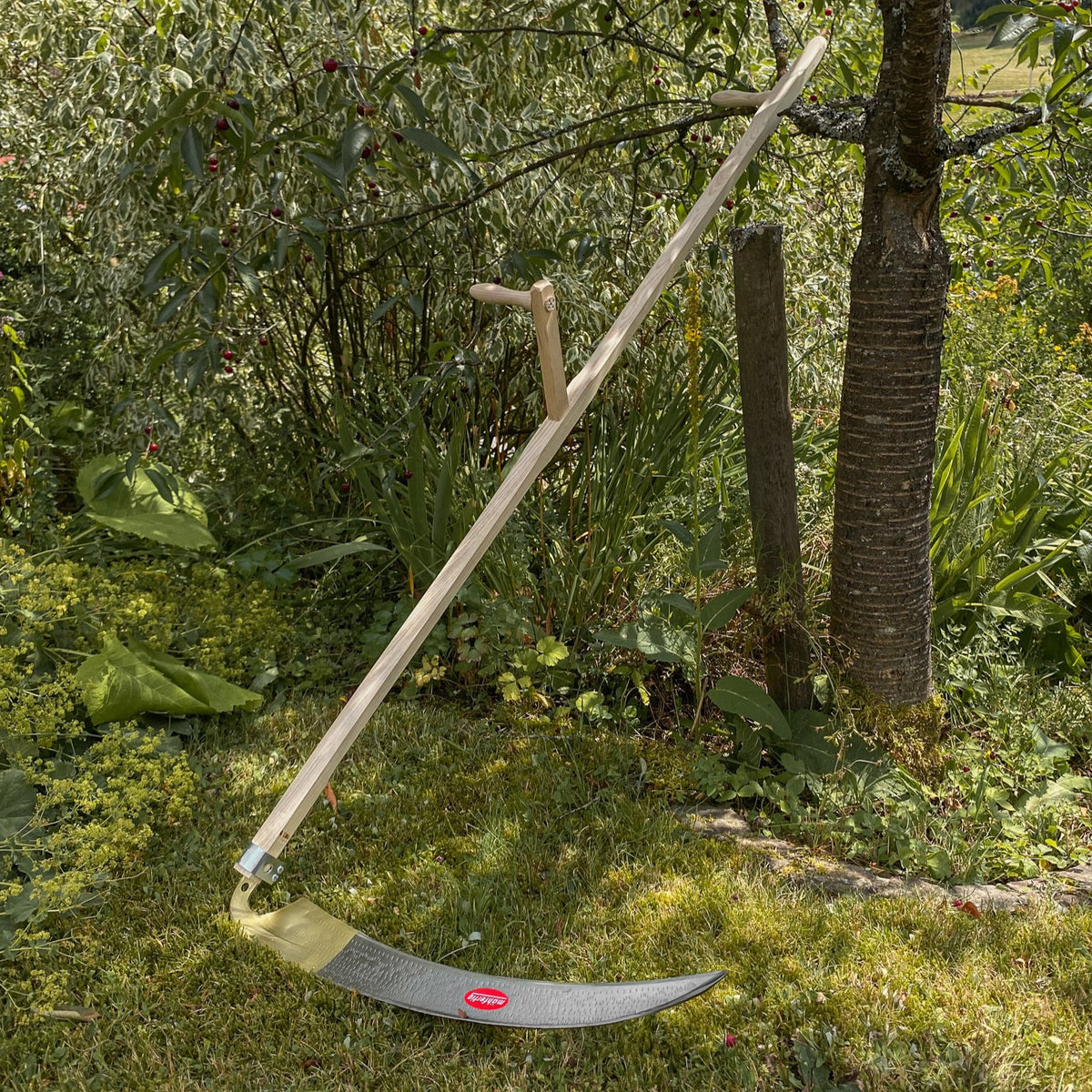 SHW Sense Friedrichstal mit traditionellem Holzworb, zwei Holzgriffen und gebogenem Sensenblatt, angelehnt an einen Baum im Garten. Handsense zum Mähen von Gras und Wiesen.