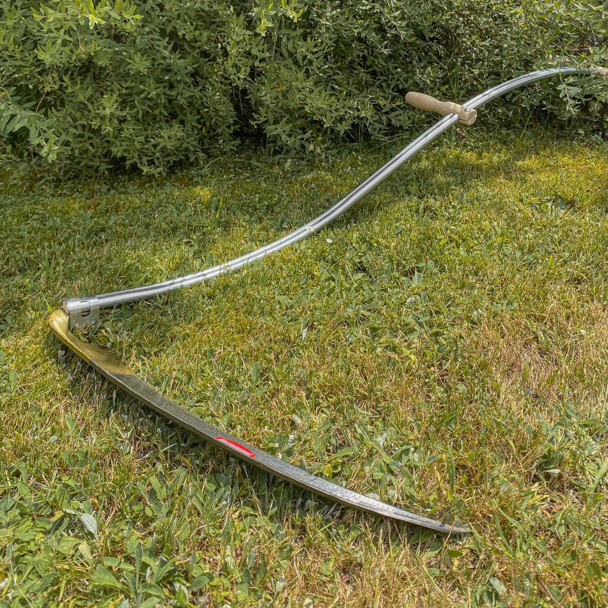 SHW Sense Friedrichstal mit gebogenem Stahlrohrworb, Holzgriff und langem Sensenblatt auf einer grünen Wiese. Handsense zum Mähen von Gras und Wiesenflächen.