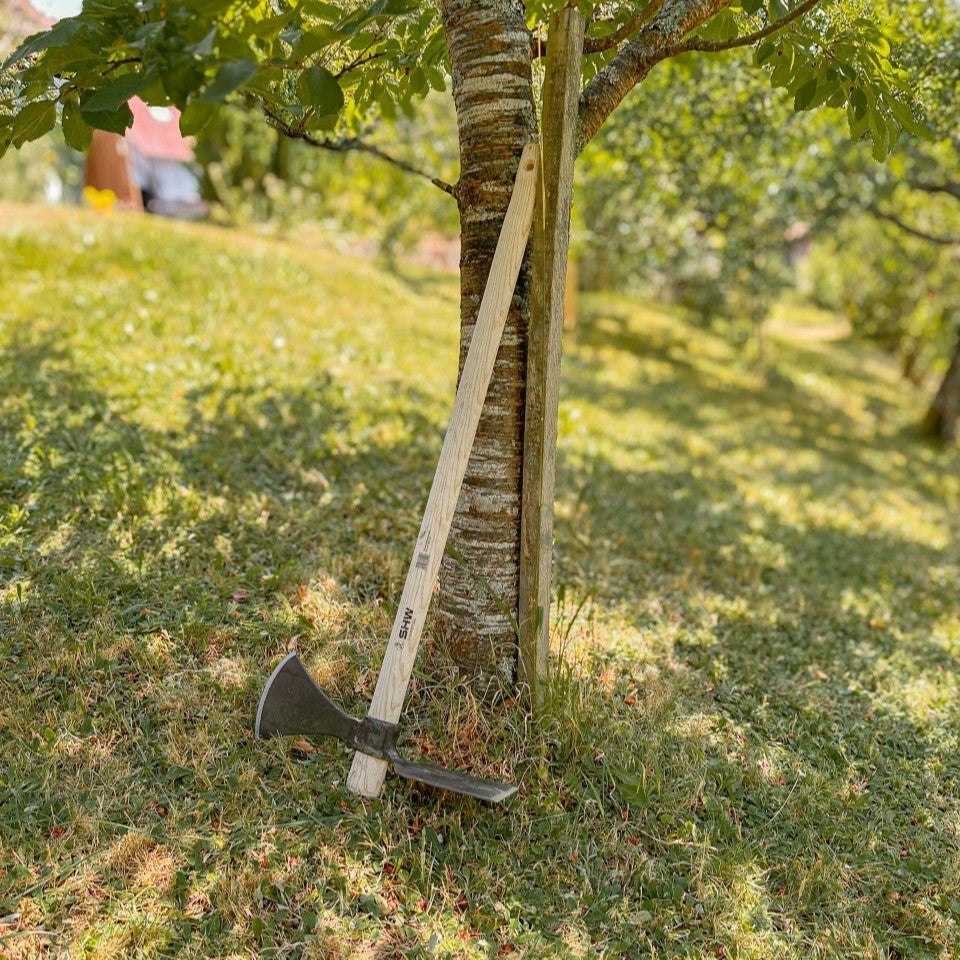 SHW Wiedehopfhacke mit geschmiedetem Stahlkopf, breitem Hackenblatt, spitzer Gegenseite und langem Holzstiel, angelehnt an einen Baum auf einer Wiese.